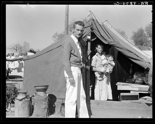 The image depicts a black-and-white photograph of three individuals, presumably from the early to mid-20th century based on their clothing and hairstyle. A man stands prominently in the foreground wearing a light-colored sweater over a dark collared shirt paired with white trousers; his expression is serious as he gazes directly at the camera.

Behind him, slightly to the right, there's an older woman who appears to be holding a baby wrapped snugly against her chest with one arm. The mother wears a long-sleeved dress and has short hair brushed back from her face, suggesting modesty in attire appropriate for daily chores or travel during that era.

On the far left stands another young girl, likely around 12-14 years old judging by her height compared to the adults; she's wearing a sleeveless blouse with long sleeves over what seems like an apron and has dark hair tied back. She appears contemplative yet somewhat resigned as she looks off into the distance.

The backdrop reveals a makeshift tent or temporary dwelling, indicative of rural life possibly on a farm where such structures were common for living quarters during that time period. Various items are scattered around outside the tents, including buckets, pots, and what seems to be laundry hanging out to dry which gives an impression of daily domestic activities being conducted amidst modest surroundings.

The atmosphere conveyed by this photo suggests hardship or dis [...]