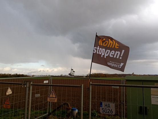 Eine schwarze Fahne mit Aufschrift "Kolhe stoppen!" (campact) wehr über einer Pumpstation am Rand des Tagebau Hambach, im Hintergrund ein Acker und am Horizont ein Schaufelradbagger von RWE