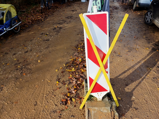 Auf einem Feldweg lehnt ein gelbes X aus Holz an einer Baustellen-Barke aus weiß-rotem Plastik, am Rand stehen PKW und ein Fahrradanhänger.
