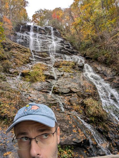 Top half of Steve's face in the lower left corner, wearing a blue Dumbo ball cap and looking up as if towards the falls. The rest of the frame is the waterfall cascading down some jagged dark gray rocks with orange and yellow leaves & trees all around. Taken at Amicalola Falls State Park in Georgia.