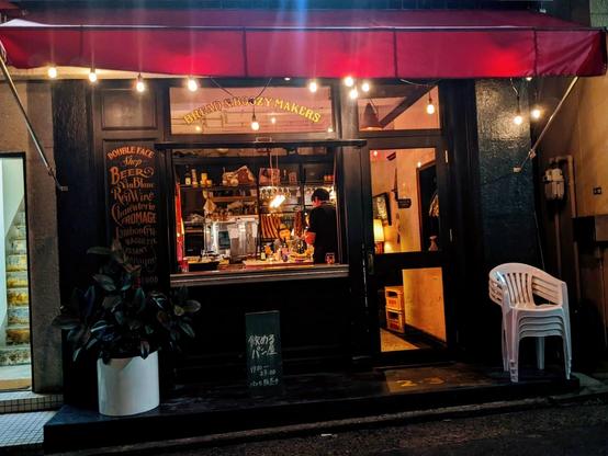 Outside of bread and Boozy makers at night, with white plastic chairs stacked and some people inside seen through glass windows