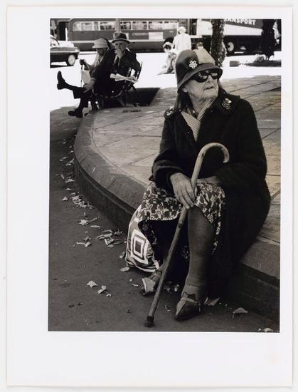 The image is a black and white photograph depicting an elderly woman sitting at what appears to be the edge of a sidewalk or curb. She's wearing sunglasses, a hat adorned with decorative pins, and has various accessories including brooches on her coat.

In front of her are scattered pieces of paper which seem like litter. The setting suggests a public space possibly in a city environment as we can observe cars passing by behind her, along with what appears to be an older style transport bus labeled 'TRANSPORT'. There's also another person seated further back and partially out of focus.

The elderly woman is holding onto a cane, suggesting she may have mobility issues. Her attire includes patterned pants or skirt, dark shoes, and seems well-fitted for cooler weather judging by her heavy coat. The overall mood of the image could be interpreted as contemplative yet somber due to its black-and-white tones and urban setting.

The photograph is credited with a source from "Cathedral Square, Christchurch" which indicates it's taken in New Zealand considering that Cathedral Square was notably affected during an earthquake there. It belongs to a series titled 'Notes on the country I live in', suggesting personal reflections or observations of daily life in this context by Ans Westra.
