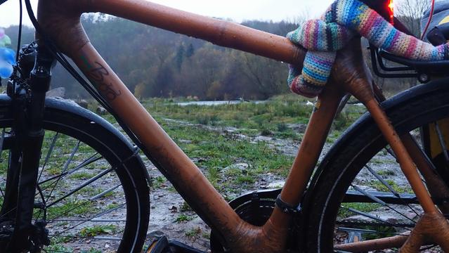 Blick durch einen Fahrradrahmen der tropfnass ist. Um die Sattelstütze ist eine Schlosskette mit buntem Hekelschlauch, gewunden