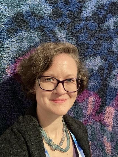 A selfie of a white, middle-aged, blue-eyed woman wearing slightly askew acrylic-framed glasses. She has short, wavy, mousey brown hair, and is smiling at the viewer. She is wearing two strings of pale blue, glass bead necklaces, a blue blouse, and dark grey sweater. She is standing in front of a thick-pile, wall-hanging rug. The rug has an abstract design in blues, greys, and violet.