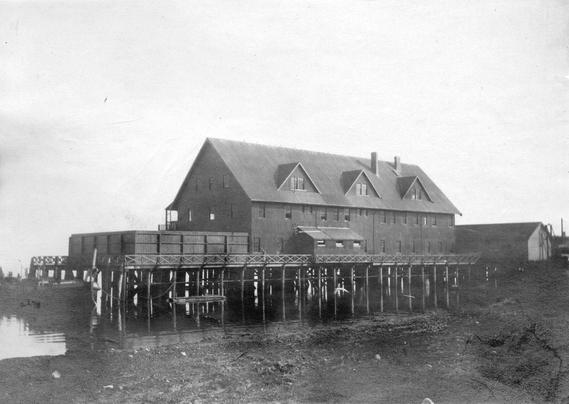 A black and white photograph of a large wooden building elevated on stilts over water, featuring a sloped roof and multiple windows. The structure has a front porch area and several wooden crates stacked alongside. The surrounding area appears flat with a muddy shoreline.