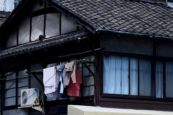 A view of the upper floor of an old Japanese-style house with light blue curtains. A clothesline with colorful clothes can be seen on the gable