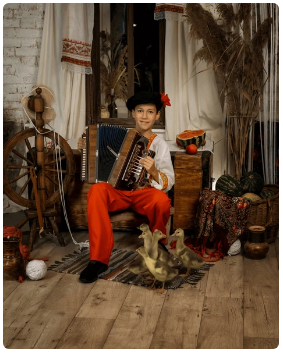 A boy dressed in traditional rural Slavic garb poses with a squeezebox in a photo studio set including a spinning wheel, a thick slice of watermelon and a small flock of ducklings.
