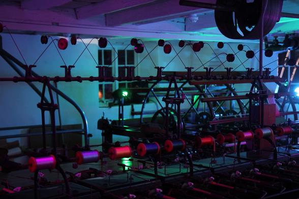 A machine for winding silk onto spools. Lit in red, blue, and purple. There is a white wall and ceiling with timber beams on the back and top. Some of the spools have silk on them in various assorted colours.