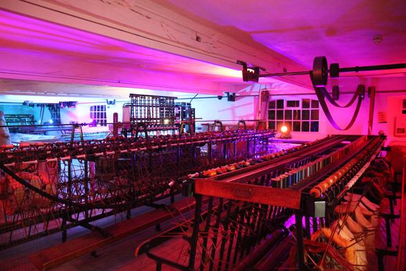 A zoomed-out image of a large room with white walls and ceilings, supported by timber beams. There are multiple different machines for the making of silk products, and the room is lit in purple, pink, blue, and white hues. The floor is lit in a deep, gentle red. All of the visible spools are empty.