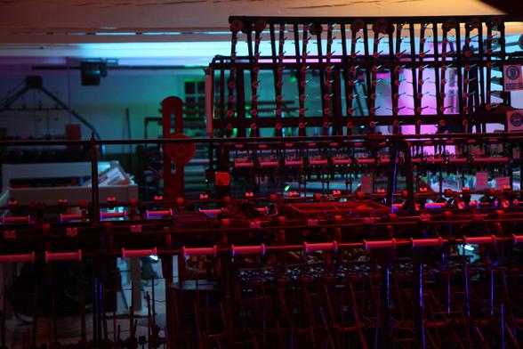 A photo of several machines for winding silk onto spools for later use. They are lit in purple, pink, and blue hues. The left side is darker on the left side, but the top of the machine is lit in a gentle red, with it getting darker towards the bottom. All of the visible spools are empty.