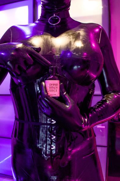 A tight cropped picture of Loki wearing a black latex catsuit and corset. 
They’re holding a bottle between their boobs labelled “Dumb Bitch Juice”.
