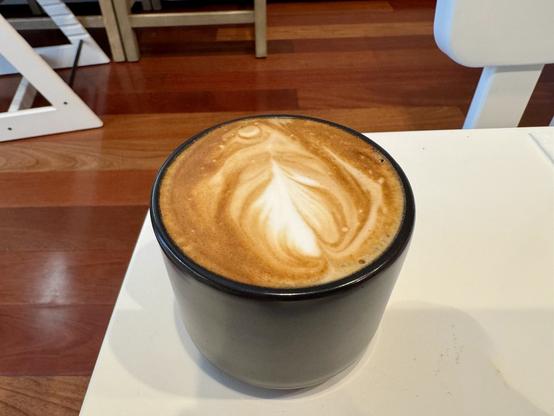Small black coffee mug filled with a cappuccino sitting on a small white table with a hardwood floor in the background.