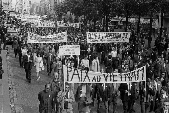 The photograph captures a high-angle view of a protest march in a Parisian street. The black-and-white image features a dense crowd of demonstrators dressed in 1960s attire, including suits and dresses. Central to the composition are large, hand-held banners displaying texts such as "PAIX au VIETNAM," "Halte à l’agression U.S.," and other messages advocating for peace. The street is flanked by trees on either side and shop fronts visible in the background, creating a sense of location and atmosphere. The overall tone is serious and resolute, embodying the spirit of activism against war.