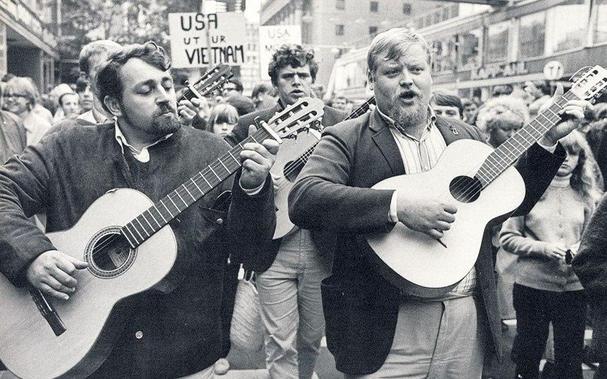 Two men, one with a beard and the other with curly hair, play acoustic guitars and sing passionately during a protest against the Vietnam War. They are surrounded by a crowd of protesters, with a sign reading "USA out of Vietnam" visible in the background. The atmosphere is lively and engaged, reflecting the historical context of anti-war activism.