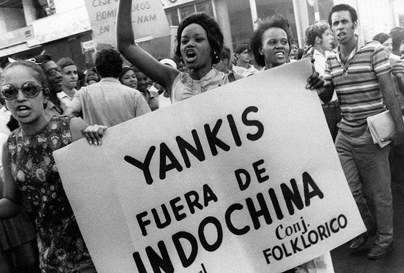 A group of protesters in a black-and-white photo demonstrates against U.S. involvement in Vietnam. Prominently featured is a Black woman holding a large sign that reads "Yankis fuera de Indochina." The crowd is diverse, with individuals expressing strong emotions. The scene captures the energy and urgency of social activism during that era.