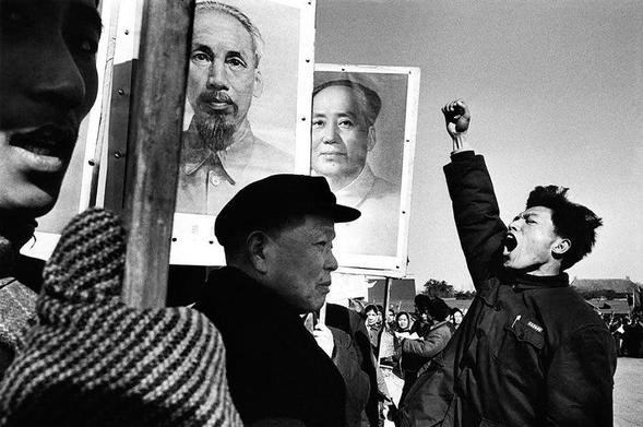 The image is a high-contrast black and white photograph. In the foreground, on the left, a young man wearing a dark jacket and shouting passionately is captured in profile, his fist raised. To his right, an older man in a black hat is depicted in a three-quarter view, looking on with a serious expression. The background features two large portrait posters of Mao Zedong and Ho Chi Minh, adding context to the scene, while a crowd of spectators is visible behind them, slightly blurred, indicating a public demonstration. The composition emphasizes the emotional intensity of the protest, with strong lines leading the viewer’s eye from the foreground to the posters.