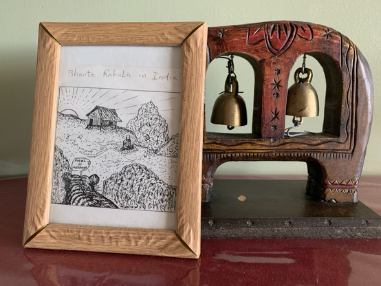 A framed drawing (in front of a wood carving with bells) of Bhante Rahula meditating in India while a tiger watches, thinking: “meal or message?”