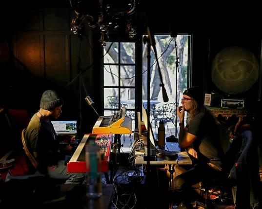 Two musicians (each wearing a black beanie) sitting face to face working on a recording together. Once musician two keyboards in front of them while the other has a desk with a large computer monitor.