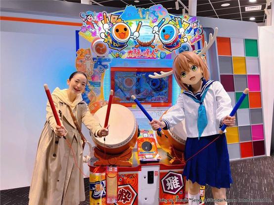 Megumi with someone cosplaying as Nokotan holding drum stick at a Taiko no Tatsujin arcade machine.