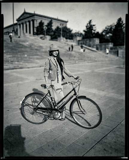 A black and white photo of a woman posing with a bicycle in front of the museum. A paper negative on 8x10.