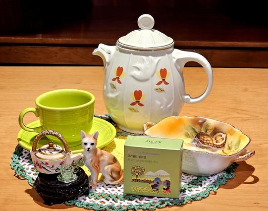 A tea tableau of a tawny cat figurine with its own tea service to mimic the larger setup for humans. There are cat-sized and human-sized versions of an orange-flowered white teapot and a bright green cup and saucer. There is a crocheted yellow and green doily, an antique painted nut dish, and a decorative green box of marigold blossom tea.