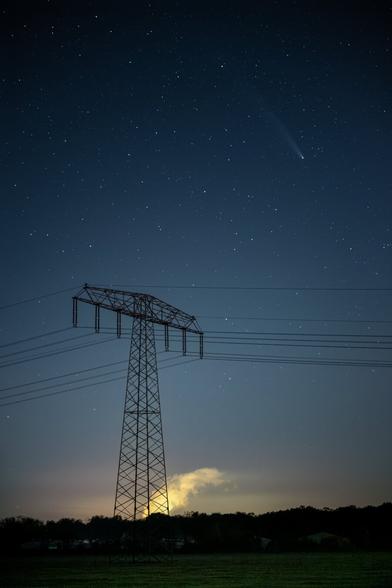 DE: Eine abendliche Hochkantaufnahme eines Starkstrom-Mastes. Links und rechts laufen Überlandleitungen auf ihn zu, im Hintergrund sieht man Lichtverschmutzung und Rauschwaden eines nahen Kraftwkeres. Und rechts oben der eigentliche "Star" der Aufnahme: Der Komet Tsuchinshan-ATLAS mit seinem langen Schweif.

EN: An evening portrait of a power pylon. Power lines run towards it on the left and right, and in the background you can see light pollution and noise from a nearby power plant. And at the top right is the real "star" of the picture: the comet Tsuchinshan-ATLAS with its long tail.