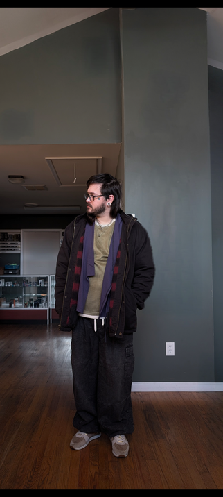 Man standing looking off to the side of the photo. He's wearing oversized black denim cargo jeans, beige shoes, a earthy green sweatshirt, with a blue kimono jacket layered on top, and an oversized black workwear jacket layered on top of that. You can see some of the black and red plaid liner of the workwear jacket peeking out. The man has on several layered necklaces, black thick rimmed eyeglasses, and long hair. There is a tattoo of a broken heart on his upper cheek next to his eye.