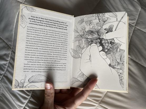 An open book with an illustrated page showing hands holding Serviceberries among foliage, featuring text discussing themes of gratitude, reciprocity, and interconnectedness in nature.