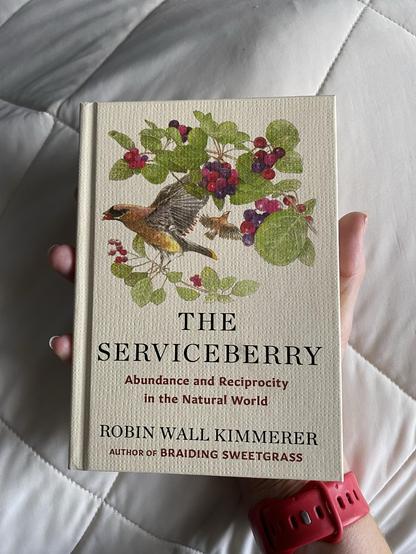 A person is holding a small hardcover book titled "The Serviceberry: Abundance and Reciprocity in the Natural World" by Robin Wall Kimmerer. The book cover features illustrations of two birds and colorful service berries surrounded by green leaves.