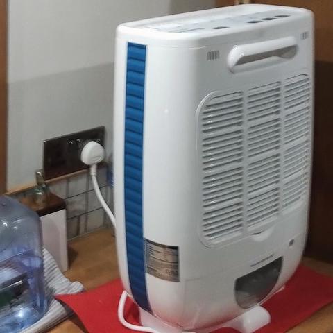 A Meaco DD8L Junior desiccant dehumidifier. It is white with a blue, ribbed stripe down the side. It has an air grille, a handle, and at the bottom, a water reservoir that pulls out like a drawer, for draining. Its white lead, or power cord, is plugged in to a 240v wall socket. The whole appliance is curvey, roughly 2 feet high, 18 inches wide, and 9 inches deep. It's sitting on the oak galley worktop on a red, foam-rubber placemat. Behind it are a soap dispenser and a refillable, clear-plastic water jug sitting on a folded tea towel. There's a bit of backsplash tiling on the wall. To the left is a bulkhead.
