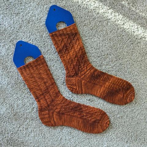 The Boulevardier Socks, knit in tonal amber, have columns of mirrored left and right twists over a stretchy ribbed background.