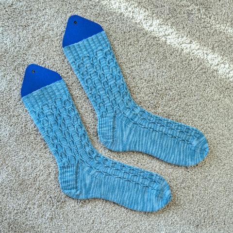 The Sapphire Martini Socks, knit in a bright blue, have staggered twists that wave back and forth over a stretchy ribbed background.