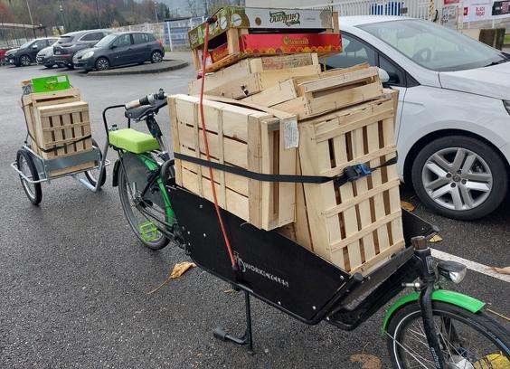 Superbe (oui c'est le mien) vélo cargo noir et vert, attelé d'une remorque et stationné sur une place de parking.
Le vélo et la remorque sont chargés de dizaines de cagettes.