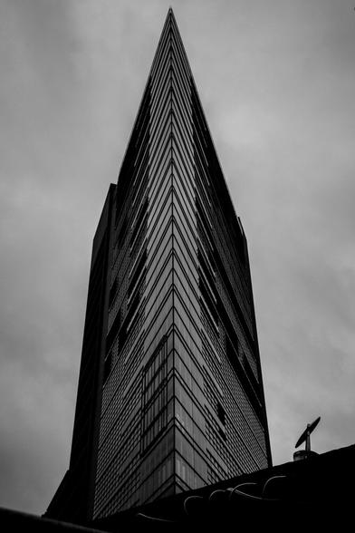 Schwarz-Weiß-Foto eine Hochhauses am Potsdamer Platz. Die Kamera schaut von unten direkt auf den spitzen Winkel der einen Kante des Hauses. Der himmel macht einen großen Teil des Bildes links und rechts vom Haus aus und is grau.