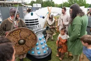 A group of people, some in historical costumes, gather around a Dalek replica outdoors. A person in green and a child look on. The atmosphere is lively and engaging.