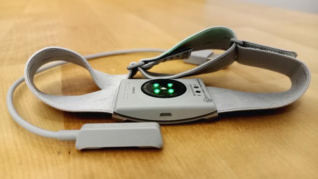 Ein grauer Heartbeat-Sensor für den Oberarm liegt inklusive USB-Ladekabel auf einem Holztisch.