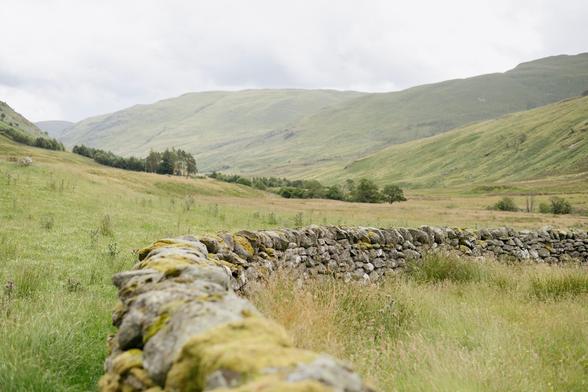 A colour photograph of a drystone sheep stell sitting in a lush green field with hills behind it.