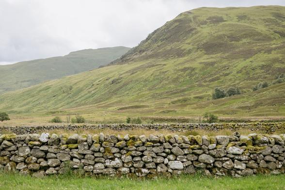 A colour photograph of a drystone sheep stell sitting in a lush green field with hills behind it.