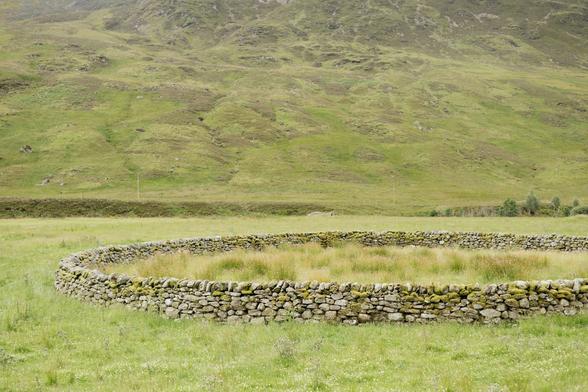 A colour photograph of a drystone sheep stell sitting in a lush green field with hills behind it.