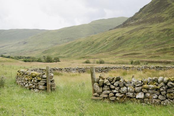 A colour photograph of a drystone sheep stell sitting in a lush green field with hills behind it.