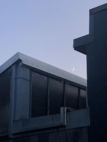 Between the pitched roofs of two factory buildings, a crescent moon on a blue purple sky.