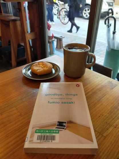Photo is of brown wooden table at angle. In front is the paperback library book with a bar code from library in the left corner. The cover is a photo of a bright corner of a minimalist room with white walls, white futon, and open computer on the floor. Above the book is a brown cookie on a moss gree plate. To the right is a grey mug of black coffee. In the distance you can see the black pants of 2 people walking past  the cafe to the left and a bunch of umbrella handles in a canister to the right