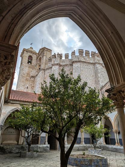 Blick durch einen angeschnittenen Spitzbogen eines Kreuzgangs durch den Innenhof mit kleinem grünen Laubbaum auf eine deutlich größere Rundkirche im Hintergrund