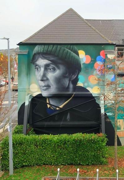 Streetartwall. A portrait of actor Cillian Murphy in his role as "Bill Furlong" in the movie "Small Things Like These" was sprayed/painted on the exterior wall of a two-story building. The mural is mainly in shades of green and shows a young man who can be seen up to his shoulders. He wears a black woolly hat and a coat and looks thoughtfully in front of him. Colorful lights can be seen in the background, which extend over a long adjacent wall. Only a part of the mural can be seen in the photo. A simple silhouette of the city is painted next to the portrait.This side will be completed in December 2024 by another artist.
Info: This new mural celebrates both the recent movie and the book (written by Claire Keegan), which tell a powerful story set in New Ross. The book and movie are set in an Irish town in 1985. In the weeks leading up to Christmas, Bill Furlong, a coal and timber merchant, is facing his busiest time. As he makes his rounds, he senses the past closing in on him - and encounters the complicit silence of a people controlled by the church.