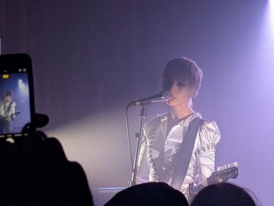 Stella Rose performing live. She is a young blonde woman, dressed in shiny white. She lays on the right side of the frame. She is in front of a microphone, singing, while playing the guitar (only the strap and the end of the mast can be seen). There are two light sources on her, one from the back (top right) and another one from the side (left). On the left side of the frame some hands are holding a phone taking a picture of the artist.