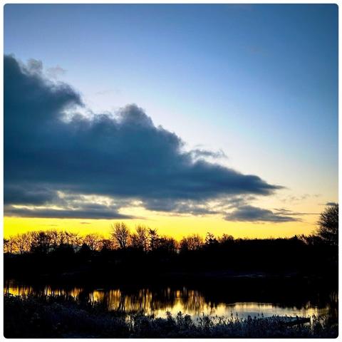 Morning sky with clouds and blue and yellow colors. In the forgeound is a lake and further behind a line of trees.