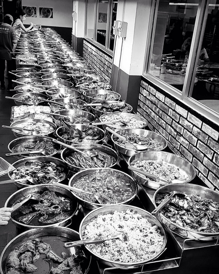 Three rows of food in trays at a self-serve economy rice restaurant. The rows are against a brick wall with large windows through which we can see the separated dining area.