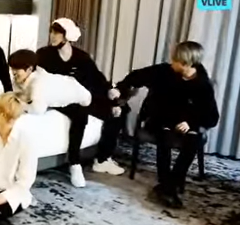 Low-resolution capture from a livestream of stray kids in a hotel room. Lee Know sits on the edge of a bed, wearing a sheeps-head towel turban, and holds Seungmin over his lap. Han sits nearby and is reaching out to move Lee Know's arm so he can get clear access to spank Seungmin's butt.