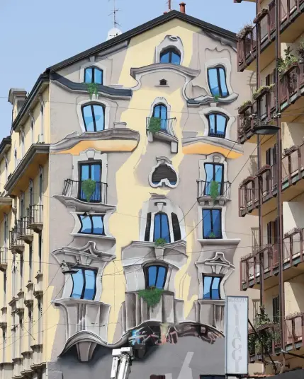 A stunning trompe-l'œil mural by Cosimo Cheone Caiffa in Milano, Italy, featuring a surreal, distorted building facade. The painted architecture appears to warp and ripple as if melted, with windows and balconies bending in impossible directions. Vibrant blue skies are reflected in the windows, while greenery spills from some of the balconies, adding a touch of realism to the fantastical design. The mural seamlessly transforms the ordinary structure into an imaginative work of art, captivating viewers with its optical illusion and creative mastery.
