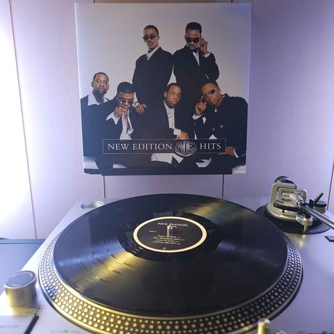 A vinyl record sits on a turntable. Behind the turntable, a vinyl album outer sleeve is displayed. The front cover shows the members of New Edition in suits, posing for the camera.